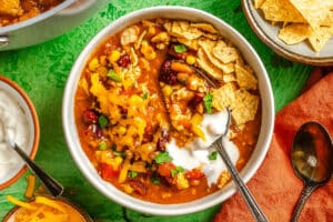 A bowl of chili topped with shredded cheddar cheese, sour cream, chopped herbs, and crushed tortilla chips, with a spoon inside. Other dishes with chips, cheese, and sour cream are nearby.