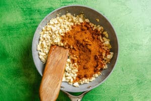 A frying pan with crumbled tofu and a mound of reddish-brown spice mixture being stirred with a wooden spatula, set on a green textured surface.