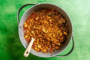 A pot of chili with crumbled tofu, beans, corn, tomatoes, and spices, on a green surface, with a wooden spoon in the pot.