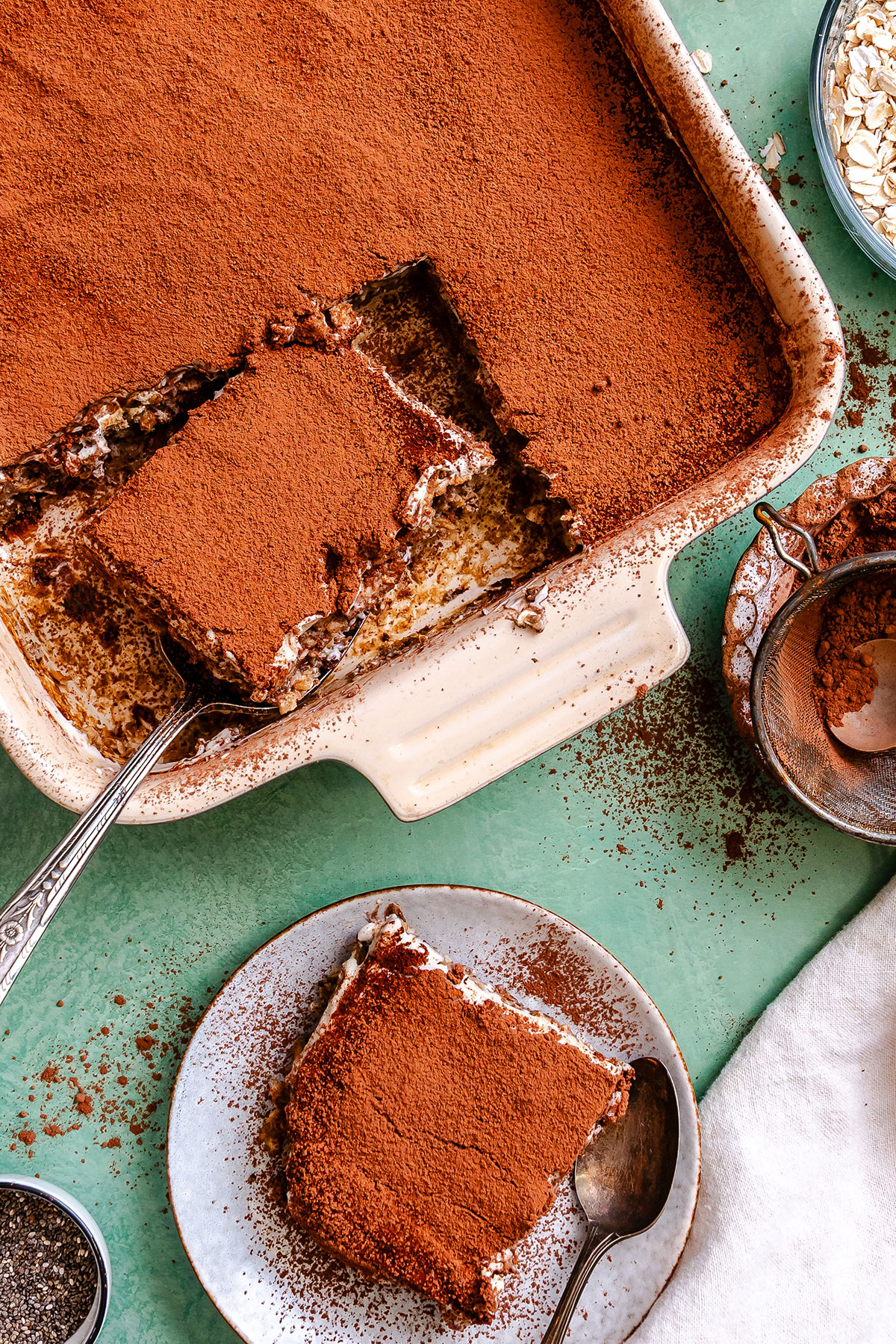 A baking dish of tiramisu with a square piece removed; the piece is served on a plate beside the dish, both dusted heavily with cocoa powder.