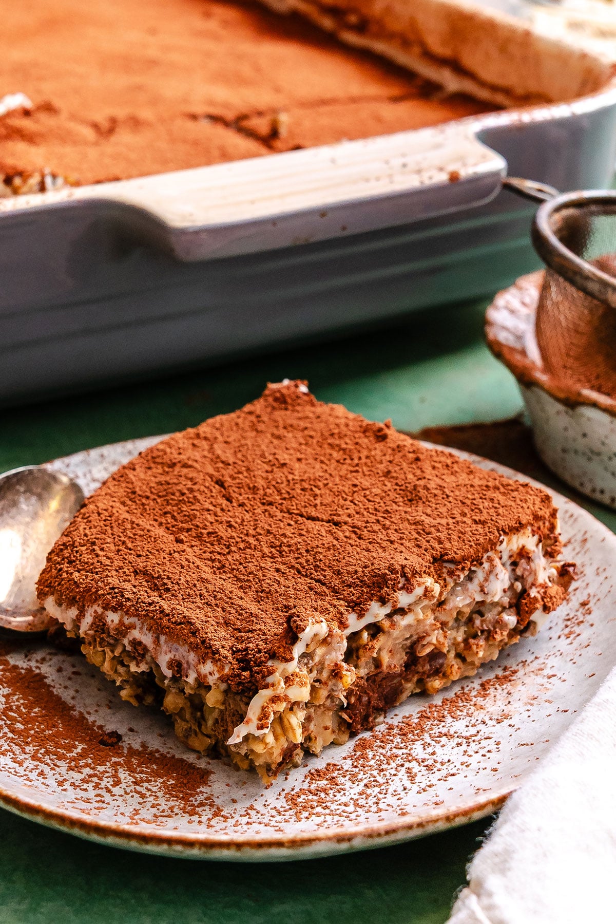 A square slice of tiramisu baked oats dusted with cocoa powder is served on a ceramic plate, with the remaining dessert visible in a baking dish in the background.