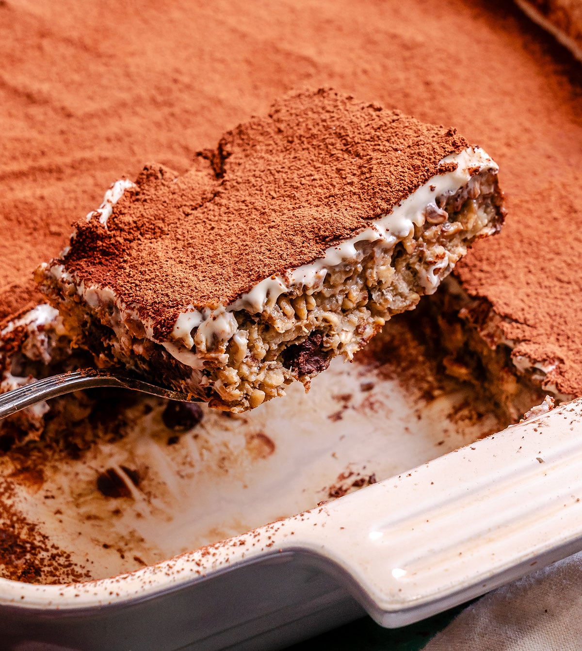 A close-up of a baked oatmeal bar being lifted from a baking dish, topped with cocoa powder and a creamy layer above an oat and chocolate base.