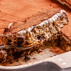A close-up of a baked oat dessert with visible nuts and chocolate, topped with cream and cocoa powder, being lifted from a rectangular baking dish.