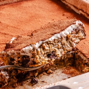 A close-up of a slice of baked oatmeal with a creamy top layer, chocolate chips, oats, and a dusting of cocoa powder being lifted from a baking dish.