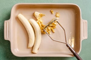 Two peeled bananas and a metal masher in a beige baking dish, with part of one banana partially mashed on a green surface.