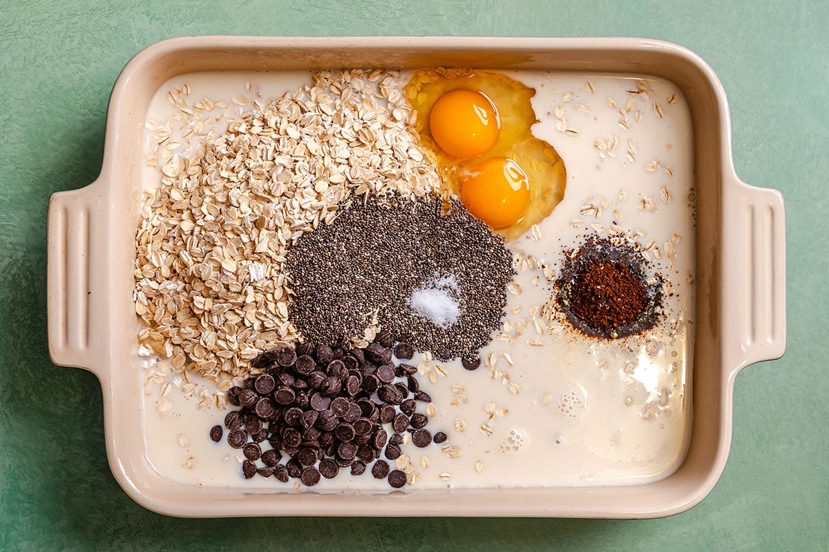 A baking dish containing oats, two eggs, chia seeds, chocolate chips, instant coffee, salt, and milk on a green surface.