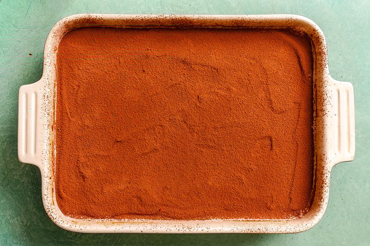Rectangular baking dish filled with a dessert topped with an even layer of cocoa powder, placed on a light green surface.