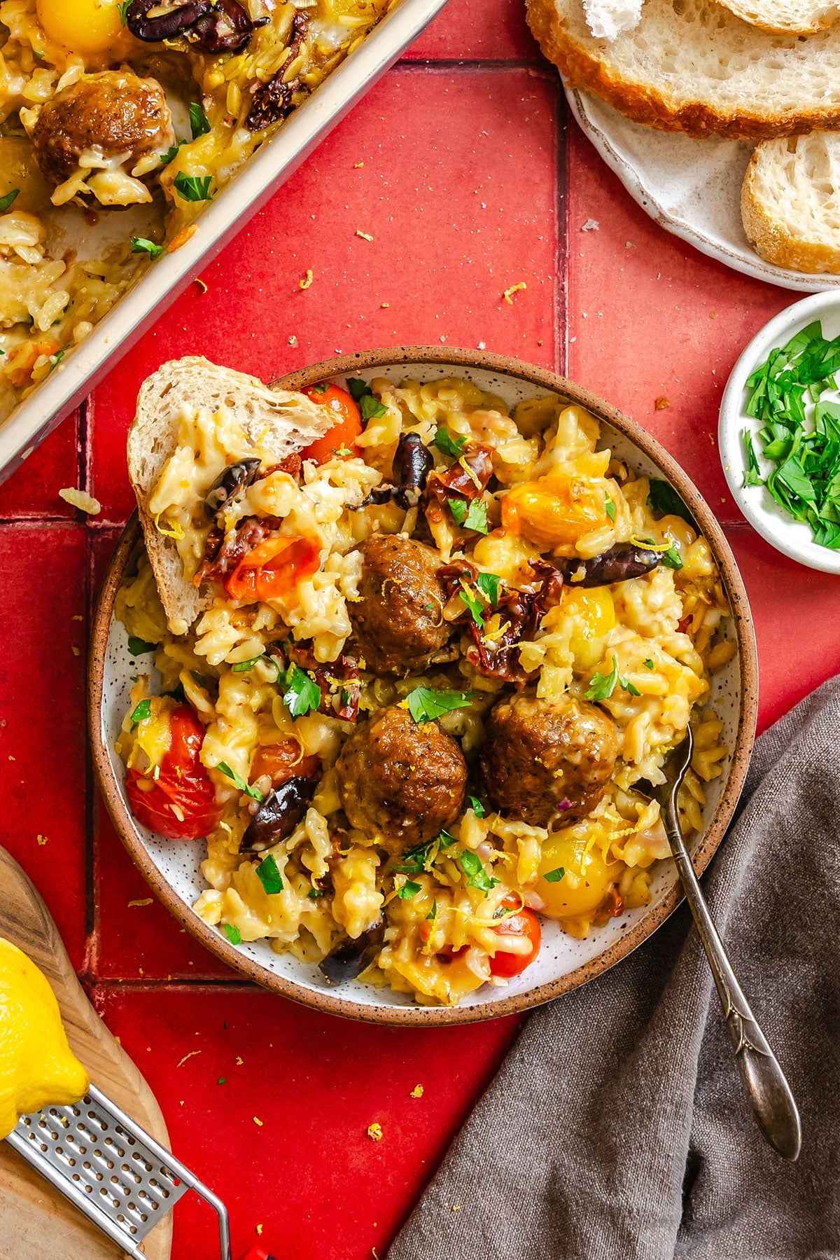 A plate of baked rice with meatballs, roasted vegetables, and herbs, served with a slice of bread on a red tile surface.