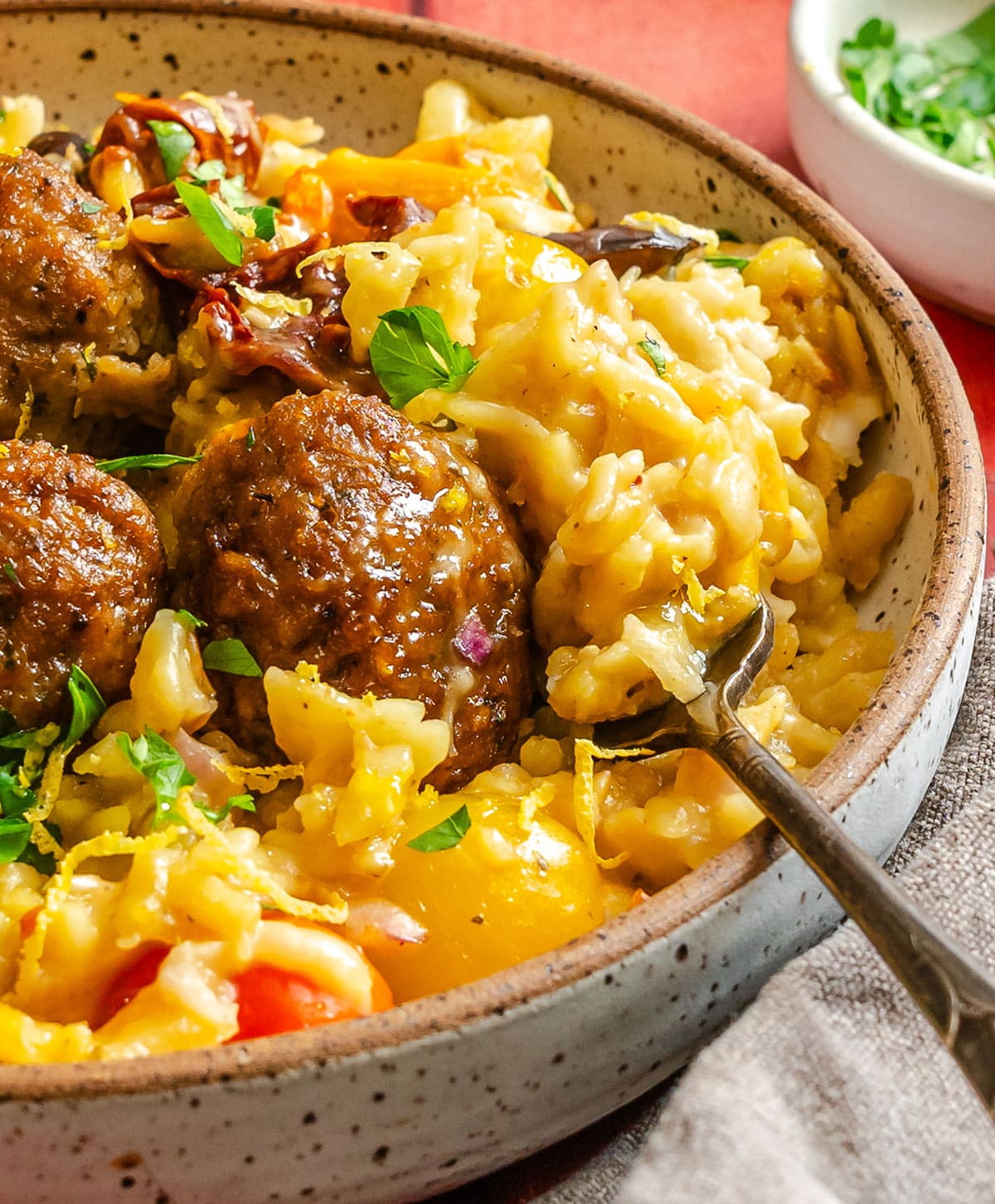 A ceramic bowl filled with orzo casserole, meatballs, and chopped herbs, with a fork resting on the side.