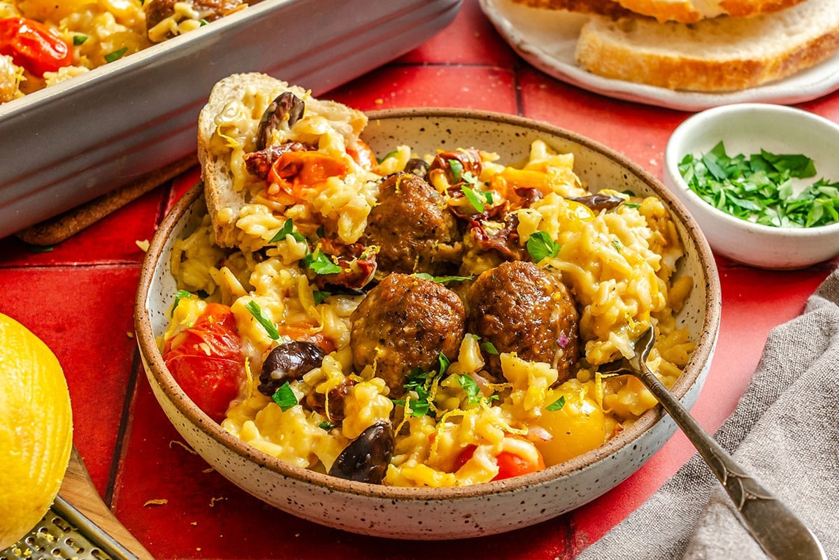 A bowl of orzo topped with meatballs, tomatoes, olives, and herbs, served with a slice of bread on a red tiled table.