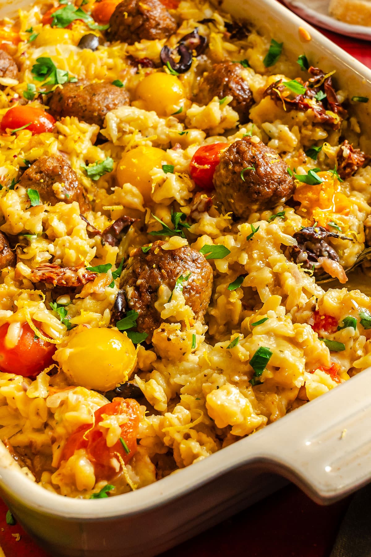 A casserole dish filled with baked rice, meatballs, cherry tomatoes, cheese, and chopped herbs.