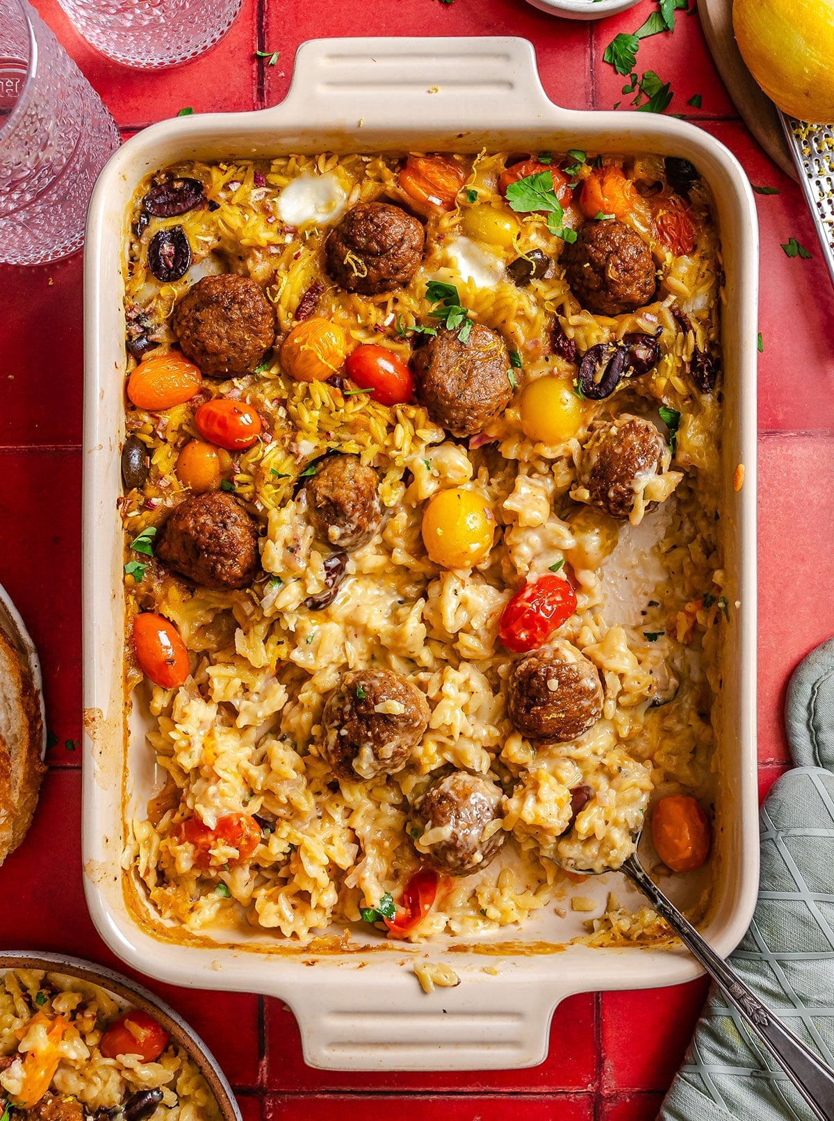 A casserole dish filled with baked orzo, meatballs, cherry tomatoes, olives, and melted cheese, with a serving missing and a spoon resting inside.