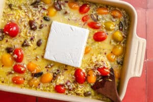 A baking dish containing uncooked orzo, cherry tomatoes, olives, chopped red onion, and a block of feta cheese, with a wooden spoon on the side.