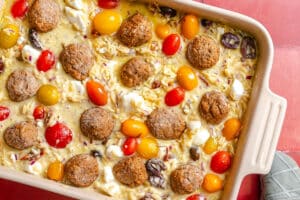 A baking dish filled with baked orzo, meatballs, cherry tomatoes, feta cheese, olives, and red onion on a red surface, with part of a shoe visible in the corner.