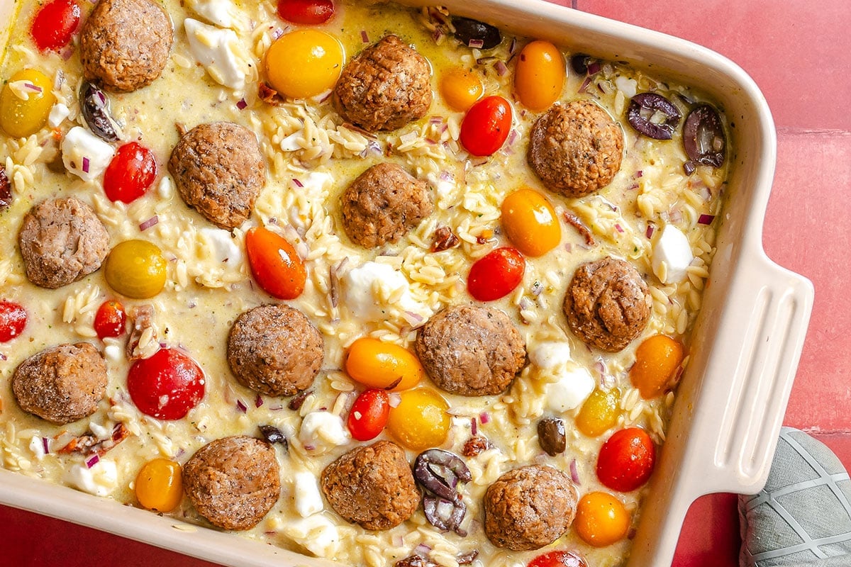 A baking dish filled with baked orzo, meatballs, cherry tomatoes, feta cheese, olives, and red onion on a red surface, with part of a shoe visible in the corner.