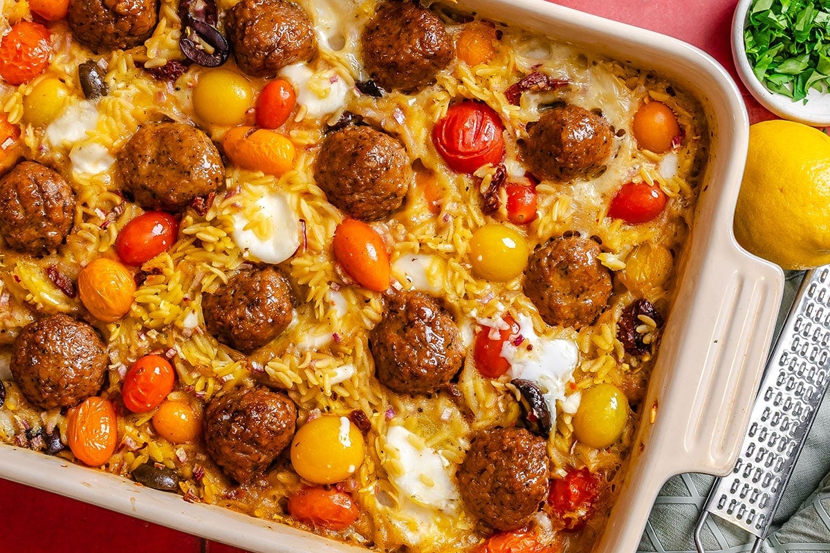 A casserole dish filled with cooked orzo, meatballs, cherry tomatoes, olives, and melted cheese, with a lemon and chopped herbs on the side.