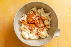 A gray mixing bowl filled with raw cauliflower florets, hot sauce, and grated cheese on a yellow surface.