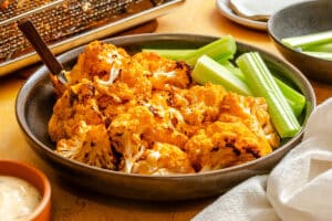 A plate of roasted buffalo cauliflower florets with celery sticks, served with a side of dipping sauce.