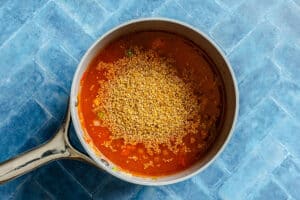 A saucepan with uncooked bulgur added to tomato-based liquid, placed on a blue tiled surface.