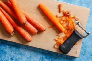 Several whole carrots and one peeled carrot with carrot peels and a vegetable peeler on a wooden cutting board, on a blue surface.