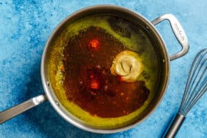 A saucepan with oil, mustard, and spices being prepared on a blue surface, with a metal whisk placed nearby.