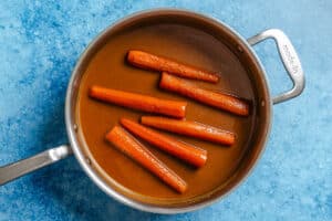 Six cooked carrots in a brown sauce inside a stainless steel pan on a blue surface.