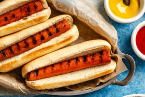 Three grilled hot dogs in buns on parchment paper, with small bowls of mustard and ketchup on the side.