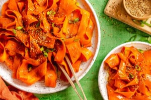 Wide carrot ribbons on plates, garnished with sesame seeds and chopped green onions, sit on a green surface next to a bowl of sesame seeds.