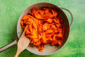 A pan filled with wide, orange carrot ribbons cooked to resemble pasta, with a wooden spatula resting on the edge, set on a green textured surface.