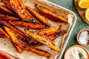 Tray of roasted sweet potato wedges sprinkled with coarse salt, served with a side of dipping sauce and lemon wedges on the side.