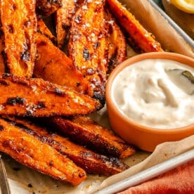 Tray of grilled sweet potato wedges sprinkled with seasoning, served with a bowl of creamy white dipping sauce and a spoon.