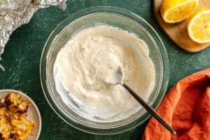 A clear bowl with a creamy white sauce being mixed by a spoon, surrounded by halved lemons, foil, a small dish of ingredients, and an orange cloth on a green surface.