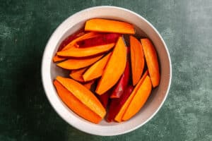 A white bowl filled with raw, orange sweet potato wedges on a dark green countertop.