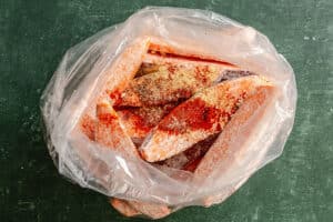 Frozen fish fillets covered in spices are placed inside a transparent plastic bag on a dark green surface.