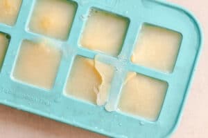 A blue silicone ice cube tray filled with frozen yellow liquid; one cube is partially broken with a piece of ice protruding.