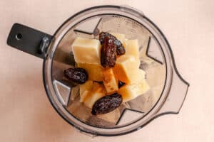 Top view of a blender containing pineapple chunks, dates, and a sprinkle of ground spice on a light surface.