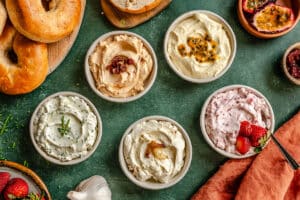Six bowls of various cream cheese spreads with different toppings are arranged on a green surface, surrounded by bagels, strawberries, garlic, and a halved passion fruit.