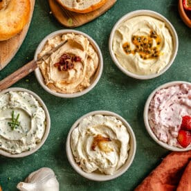 Six bowls of assorted creamy spreads, including chive, sundried tomato, passion fruit, and berry, are arranged on a green surface with bagels, strawberries, and garlic nearby.