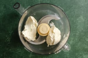 Cream cheese in a food processor bowl on a green textured surface, ready for mixing.