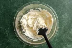 A glass bowl of thick, creamy white mixture is being stirred with a black spatula, set on a dark green countertop.