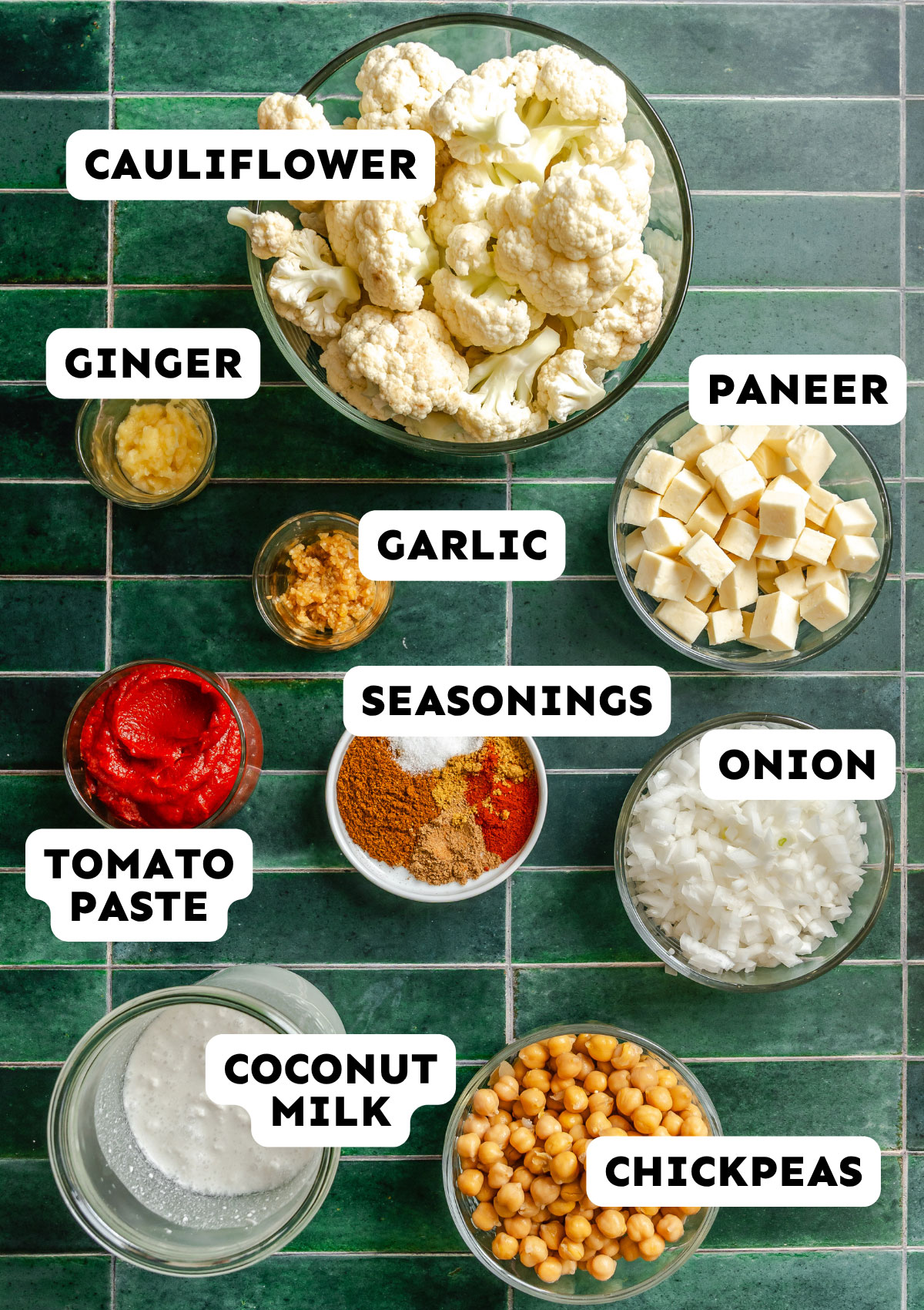 Various ingredients in bowls on a green tiled surface, including cauliflower, paneer, onion, chickpeas, coconut milk, tomato paste, garlic, ginger, and seasonings.