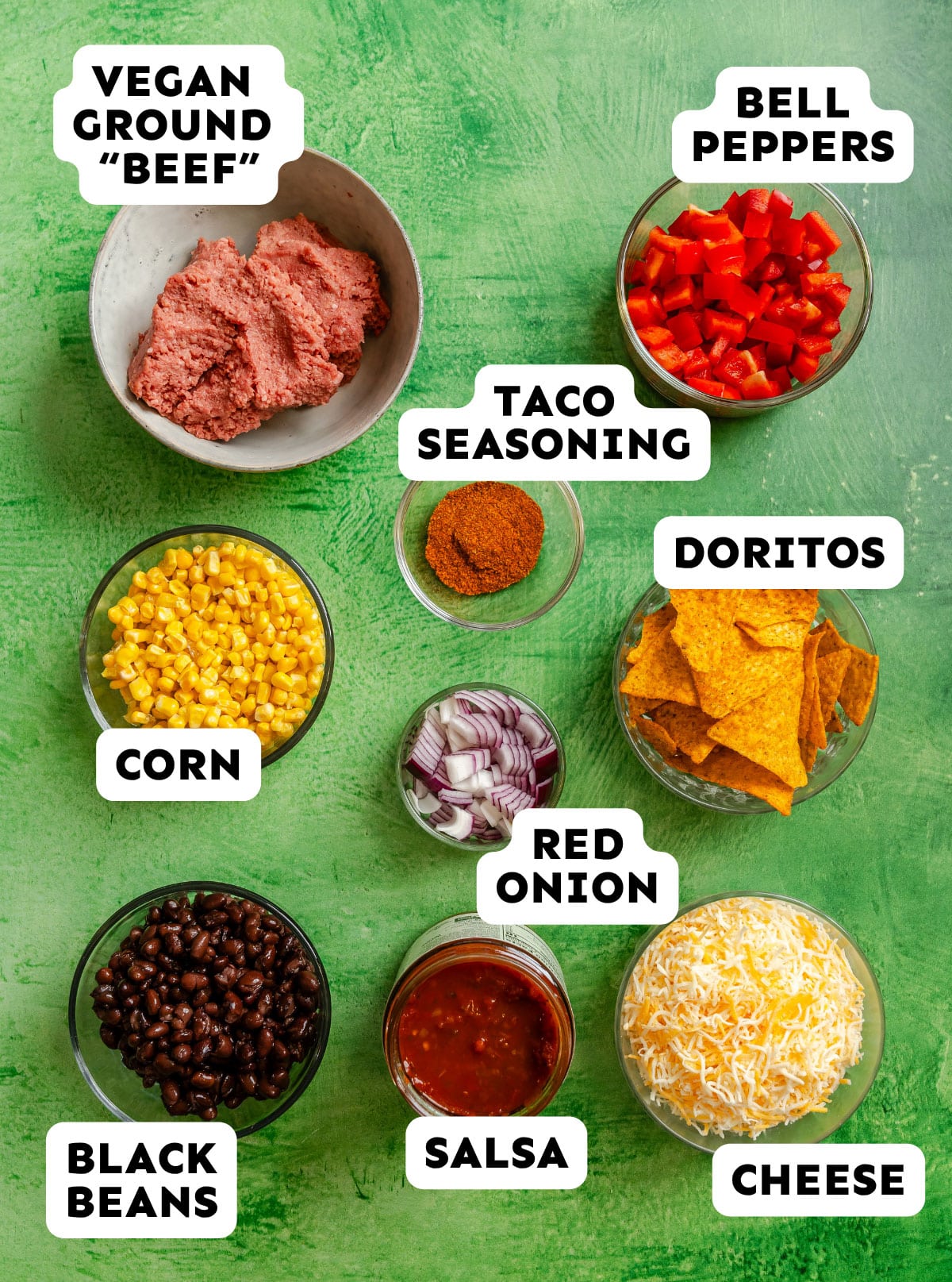 Various taco ingredients including vegan ground beef, diced bell peppers, taco seasoning, corn, Doritos, black beans, red onion, salsa, and shredded cheese on a green surface.