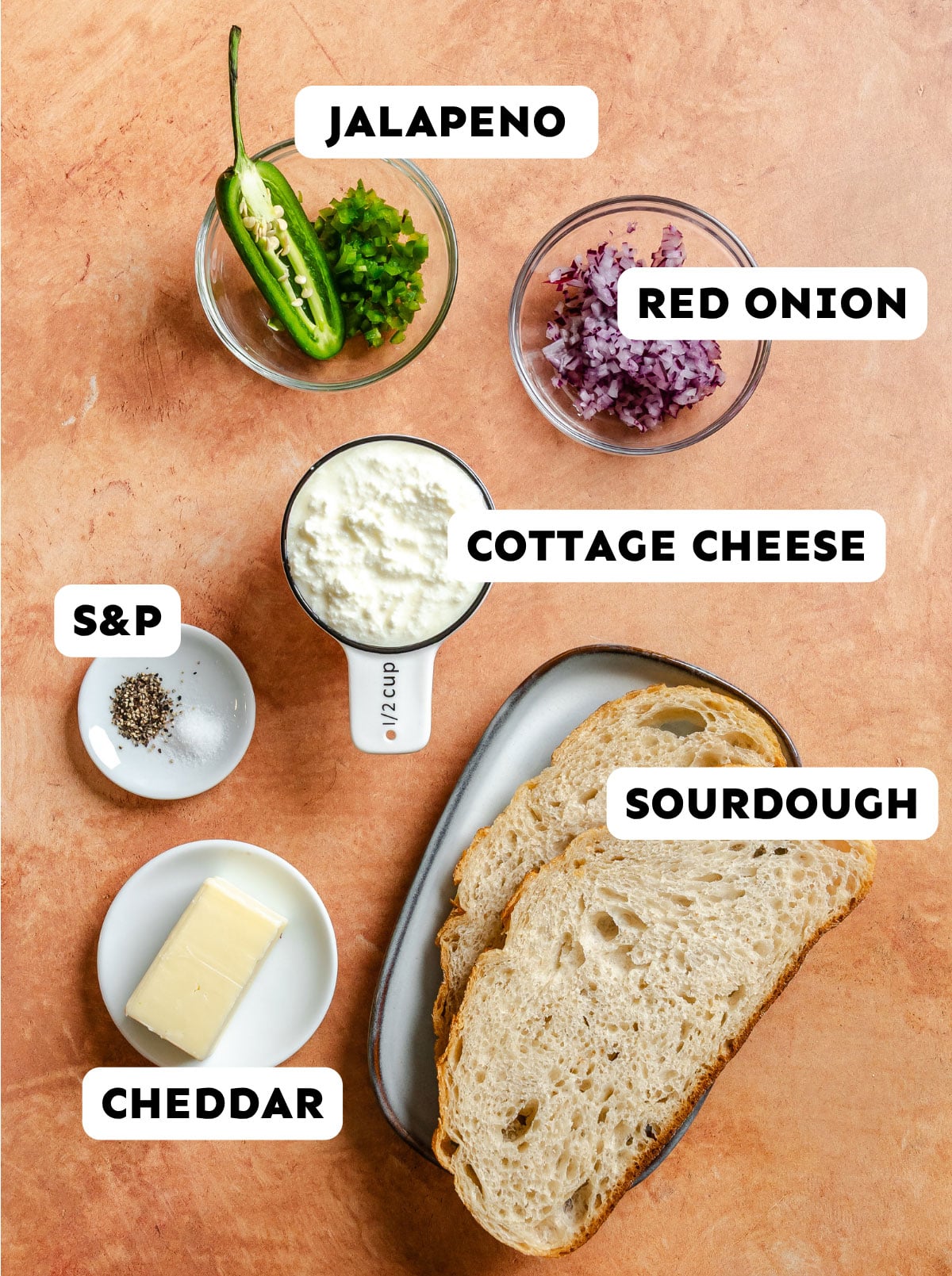 Ingredients for a sandwich on a brown surface: sliced jalapeno, diced red onion, cottage cheese, sourdough bread, cheddar cheese, and salt and pepper.