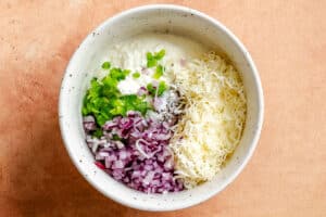 A bowl containing chopped green pepper, red onion, shredded cheese, cottage cheese, and seasonings—perfect toppings for cottage cheese toast—sits on a light brown surface.