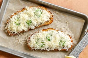 Two slices of toast topped with a white creamy spread, fresh basil leaves, and a generous amount of grated cheese on a parchment-lined baking sheet.