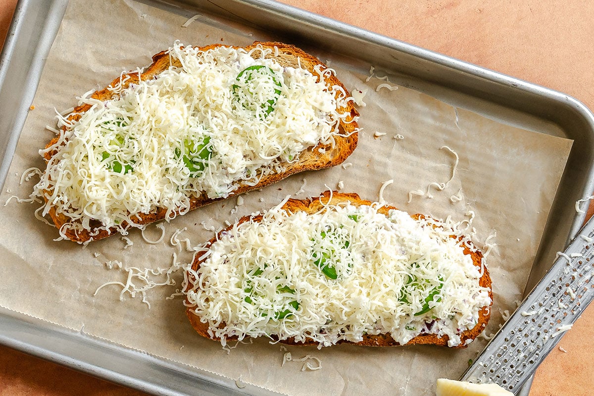 Two slices of toast topped with a white creamy spread, fresh basil leaves, and a generous amount of grated cheese on a parchment-lined baking sheet.