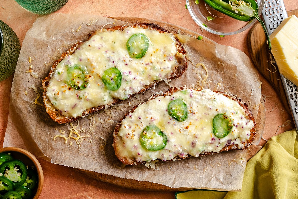 Two slices of toasted bread topped with melted cheese, sliced jalapeños, and minced red onions on parchment paper.