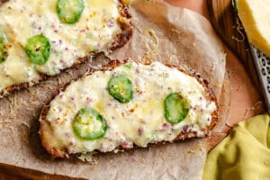Two slices of toasted bread topped with melted cheese, jalapeño slices, and bits of red onion on parchment paper.