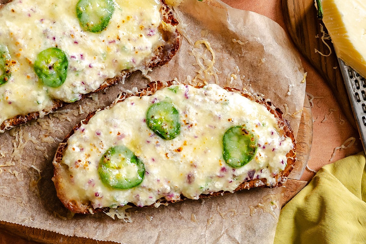 Two slices of toasted bread topped with melted cheese, jalapeño slices, and bits of red onion on parchment paper.