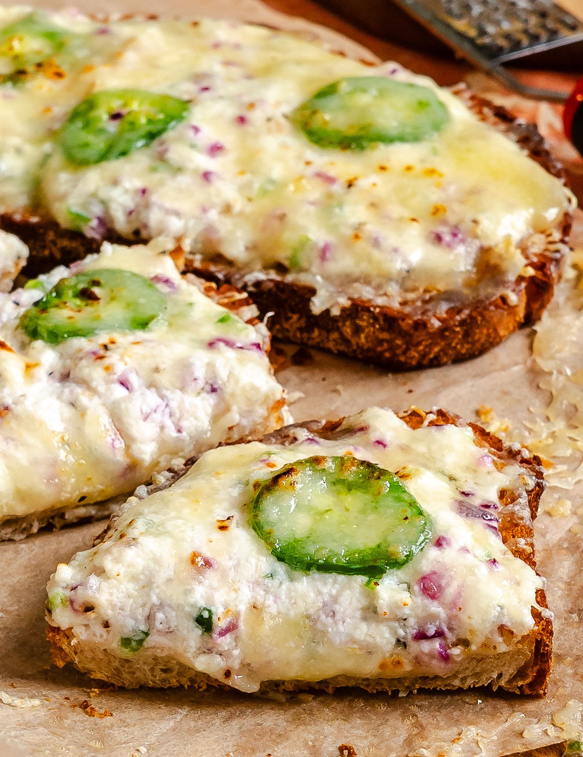 Slices of bread topped with melted cheese, chopped red onions, and sliced green jalapeños on parchment paper.